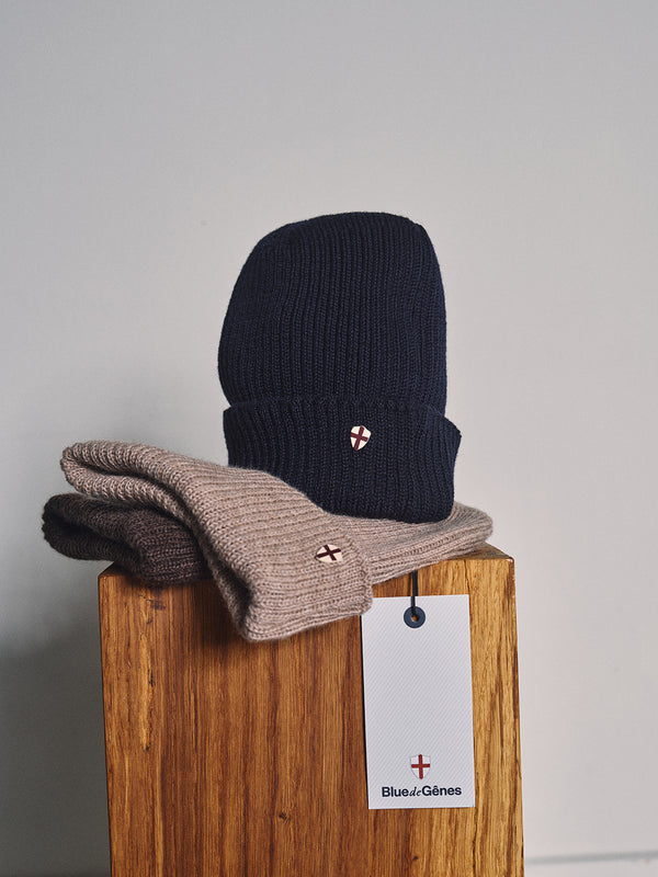A Boshi Beanie in dark navy Merino wool and a beige scarf, both with an embroidered white shield and red cross, are folded on a wooden stool beside a Blue de Gênes tag. Both pieces are expertly knitted in Denmark.