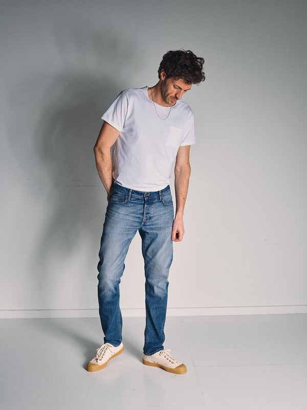 A man stands indoors against a light gray wall, looking down with his hands in the Vinci Girisha Used Jeans by Blue de Gênes. He wears a white t-shirt, beige sneakers, and soft lighting casts a gentle shadow behind him.