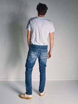 A man faces away from the camera, wearing a white t-shirt and Blue de Gênes Vinci Girisha Used Jeans—crafted from comfort stretch denim—paired with beige sneakers, standing in a plain room with white walls and a light floor.