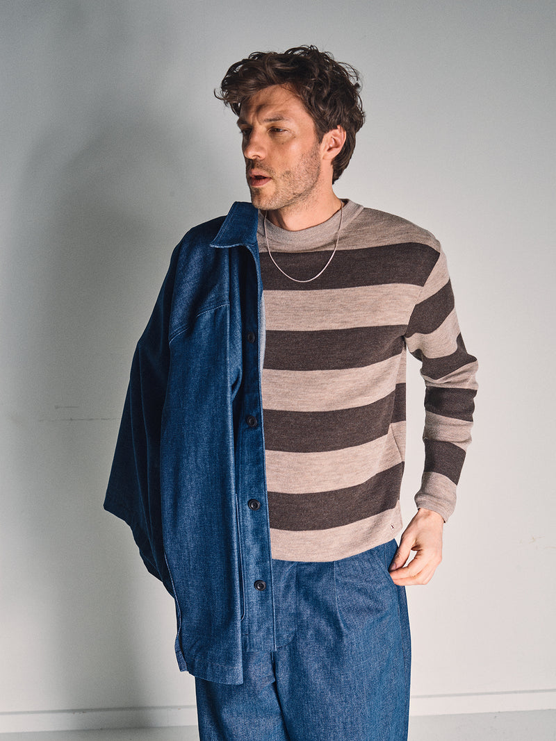 A man stands against a plain background, wearing the Blue de Gênes Toyama Stripe Knit L/S - Oat draped over one shoulder, a blue denim shirt, and blue pants. He looks to the side with a relaxed expression.