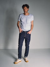 A man stands in a white room wearing a white tee, Vinci Girisha Rinse Jeans by Blue de Gênes in rinse wash, and white sneakers with tan soles. He has one hand in his pocket and looks off to the side with a relaxed expression.