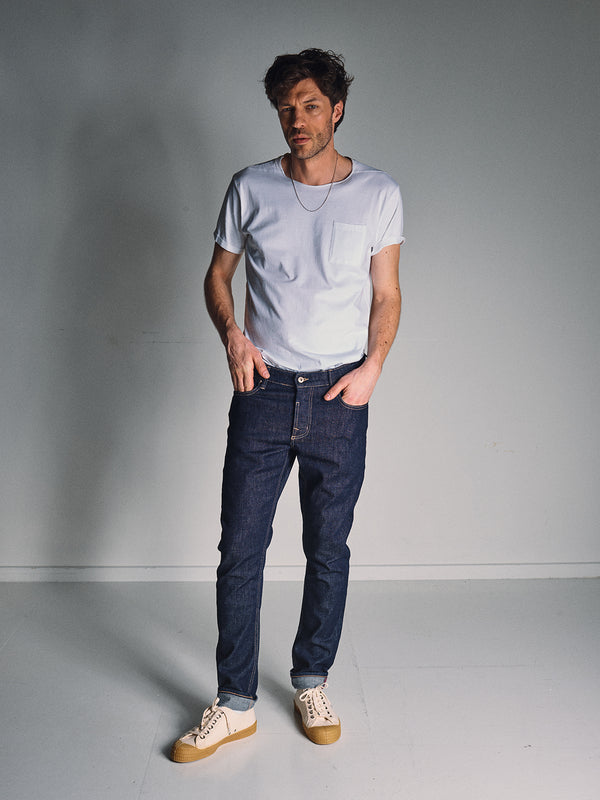 A man stands in a white room wearing a white tee, Vinci Girisha Rinse Jeans by Blue de Gênes in rinse wash, and white sneakers with tan soles. He has one hand in his pocket and looks off to the side with a relaxed expression.