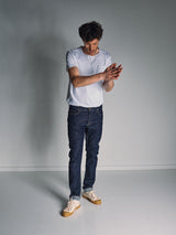A man in a white t-shirt, beige sneakers, and Blue de Gênes Vinci Girisha Rinse Jeans (Rinse Wash) stands in a minimally lit, plain room, looking down while rubbing his hands together.