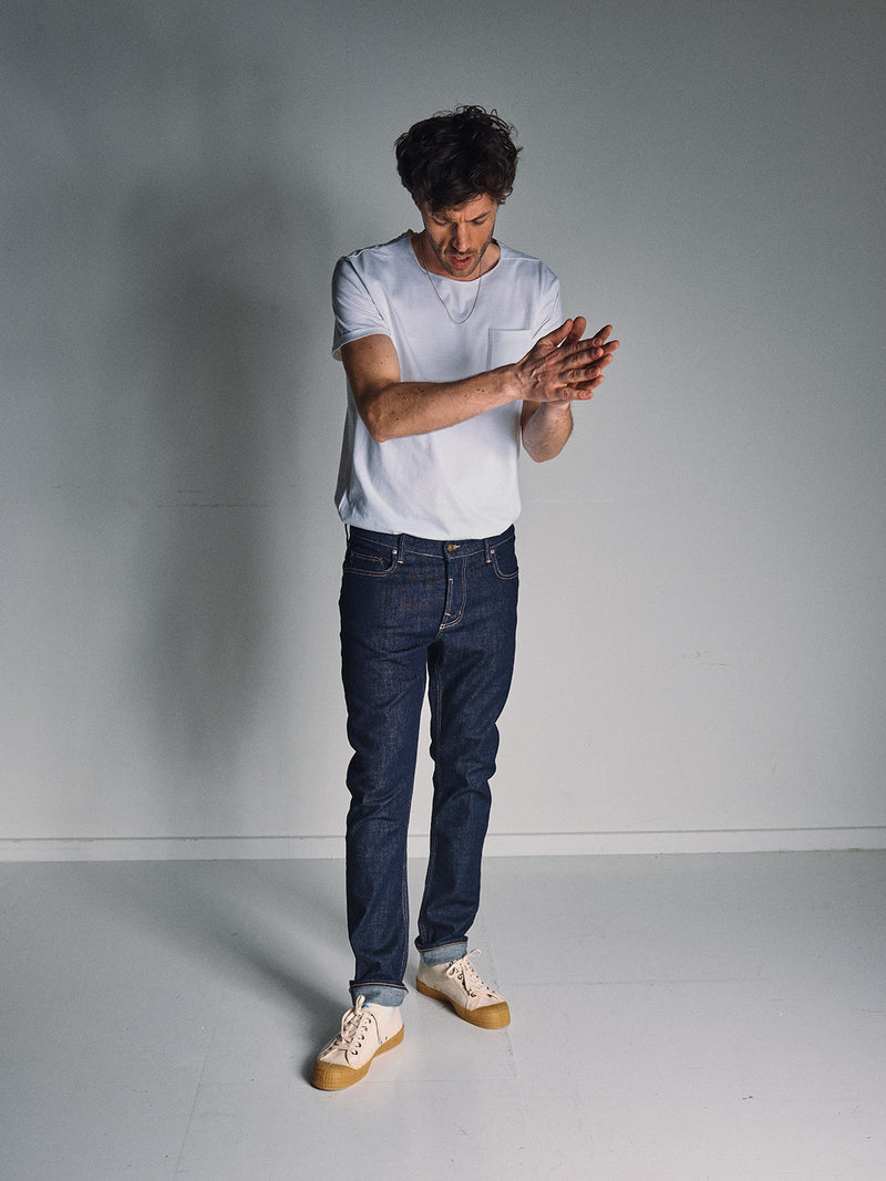 A man in a white t-shirt, beige sneakers, and Blue de Gênes Vinci Girisha Rinse Jeans (Rinse Wash) stands in a minimally lit, plain room, looking down while rubbing his hands together.