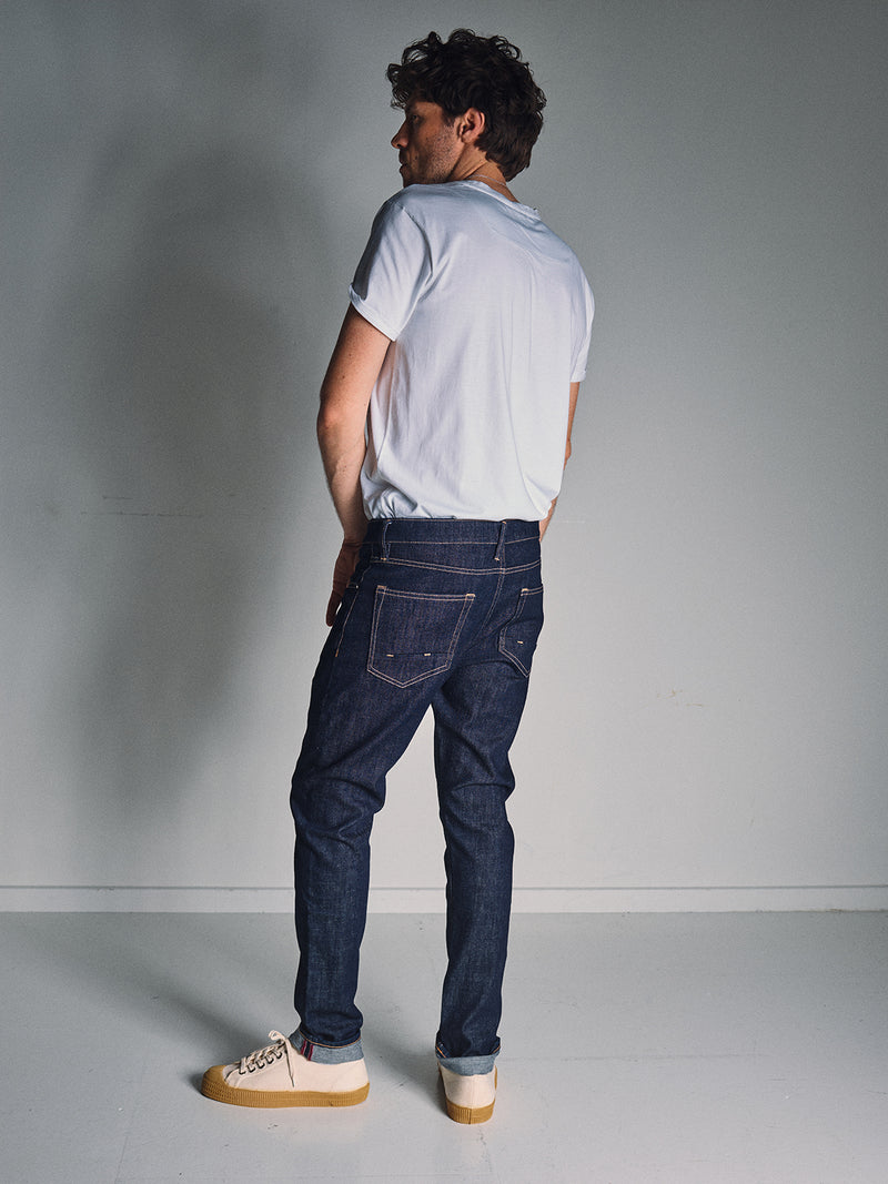 A man stands in a plain room, wearing a white t-shirt, beige sneakers, and Blue de Gênes Vinci Girisha Rinse Jeans in rinse wash. He looks over his shoulder as soft lighting casts a gentle shadow on the wall behind him.