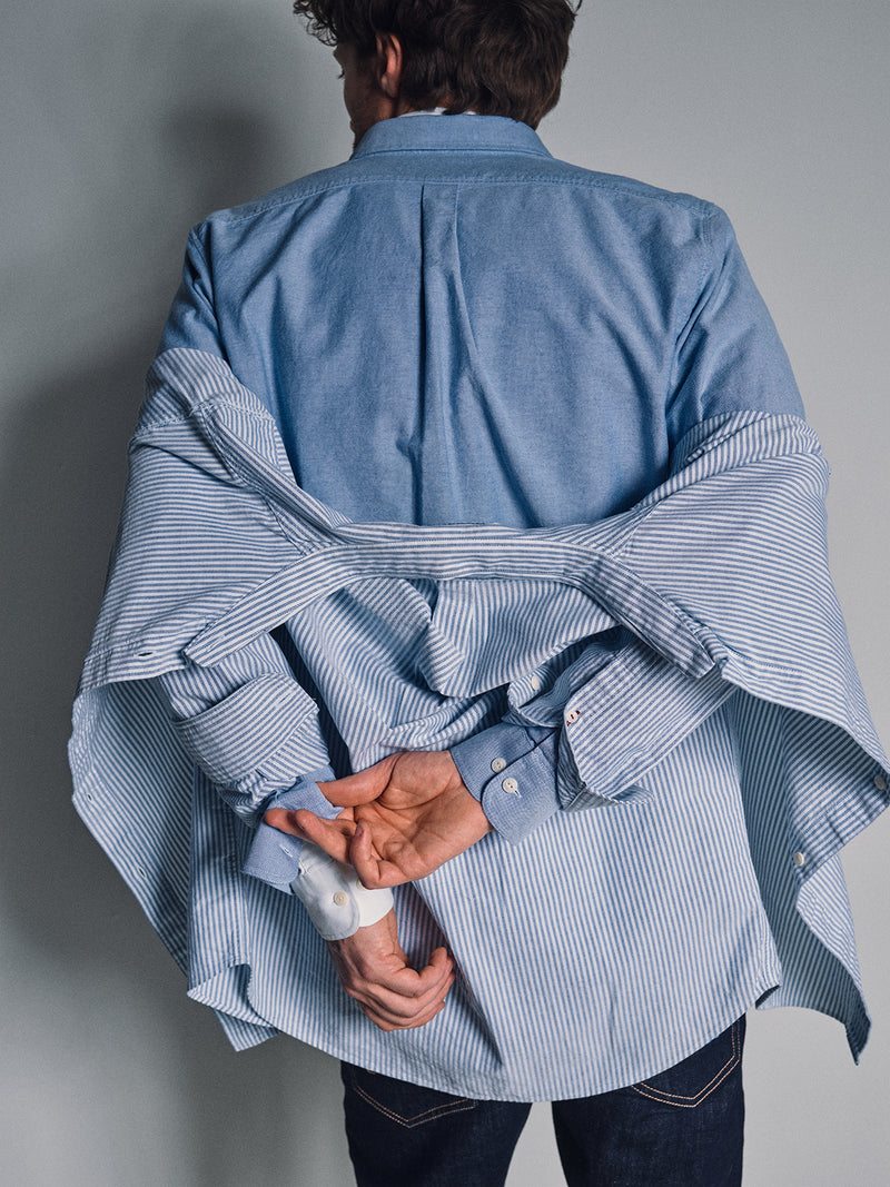 A person seen from behind wears the Sandro Oxford Shirt in blue by Blue de Gênes over a striped shirt, hands crossed behind their back and fingers holding the striped layer, against a plain gray background.