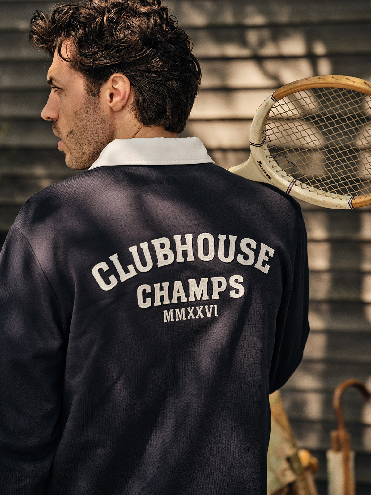A man wears the BGclubhouse Rugby Sweatshirt in navy by Blue de Gênes from the Clubhouse Champs Collection, standing with a vintage tennis racket. 