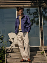 A man stands on wooden steps outdoors, wearing Blue de Gênes BGpalco Twill Pants in Chalk, a blue jacket, striped shirt, and brown shoes. Behind him are a white chair, greenery, and large glass windows reflecting trees.