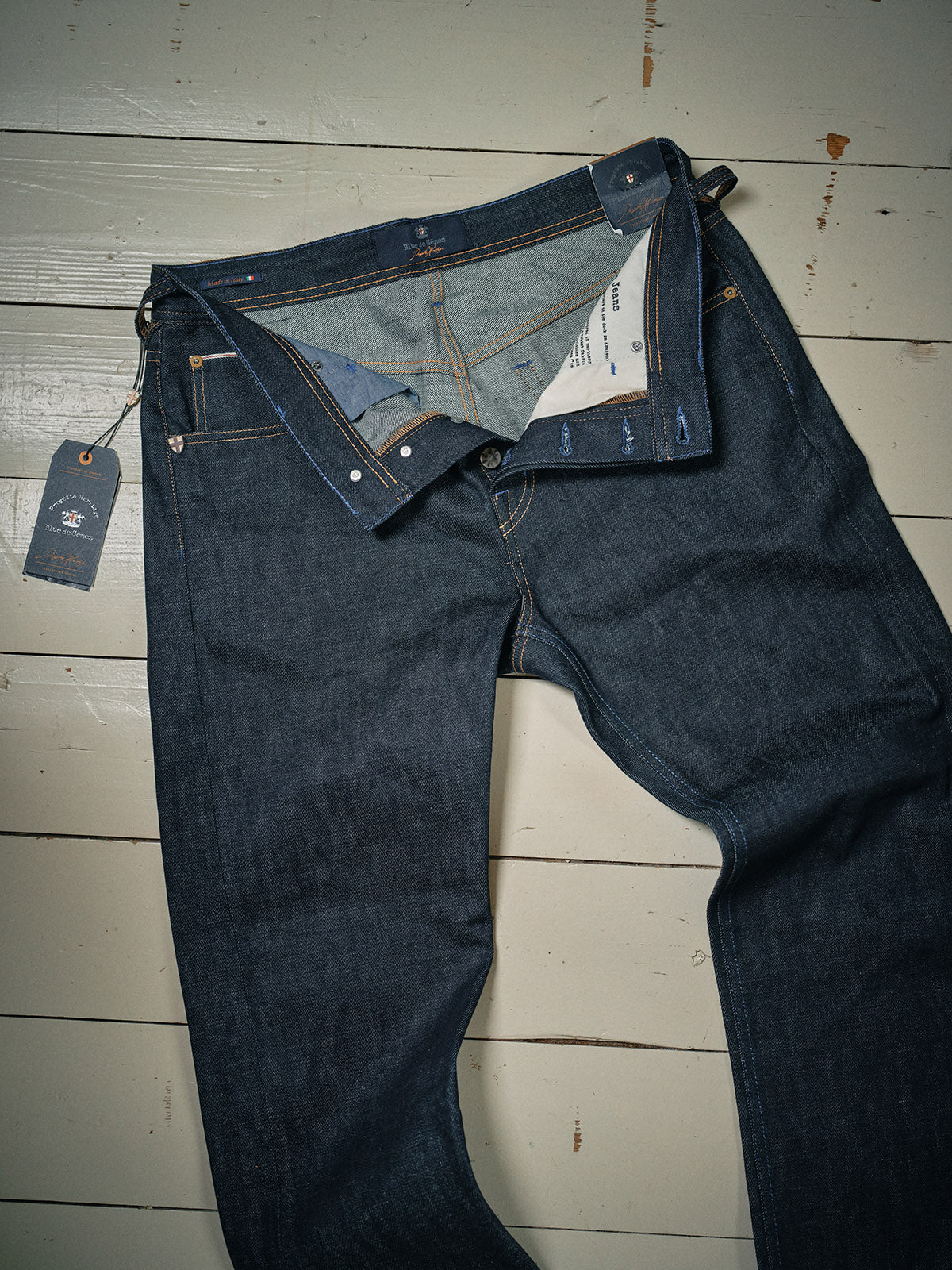 A pair of Blue de Gênes Giacomo Selvedge V2 Jeans in raw denim lies flat on a wooden floor, unbuttoned and partially open to reveal the inside. A tag is attached to a left-side belt loop.