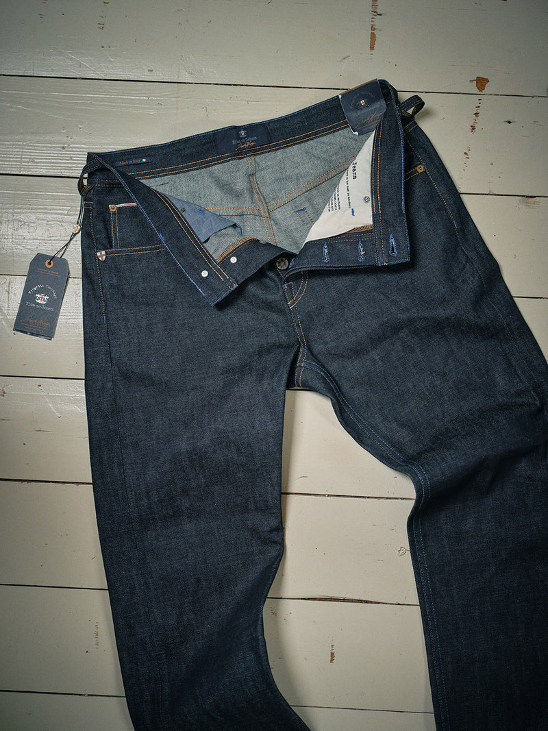 A pair of Blue de Gênes Giacomo Selvedge V2 Jeans in raw denim lies flat on a wooden floor, unbuttoned and partially open to reveal the inside. A tag is attached to a left-side belt loop.