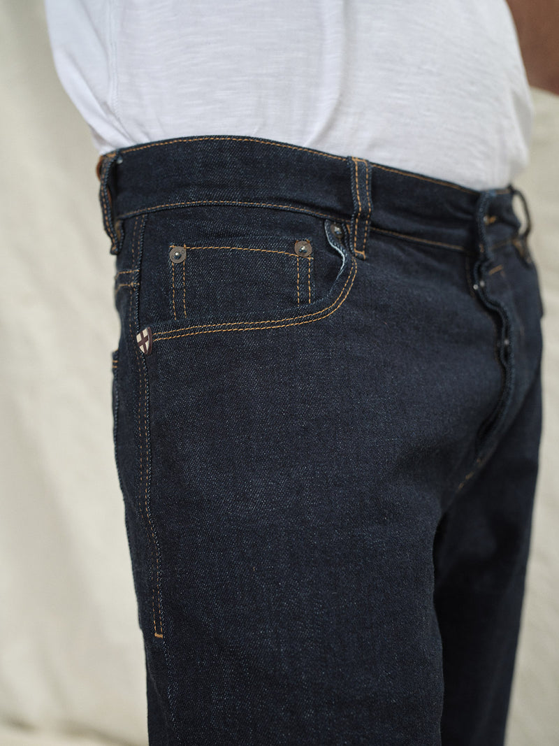 A close-up side view shows a person in Blue de Gênes Recco F V2 Jeans - Rinse Wash and a white shirt, highlighting the jeans visible stitching and pocket details against a light, blurred background.