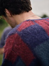 A person is seen from behind wearing the Blue de Gênes Block Knit Cardigan in a navy pattern. Their short, dark hair and a thin chain necklace stand out against a blurred outdoor backdrop.
