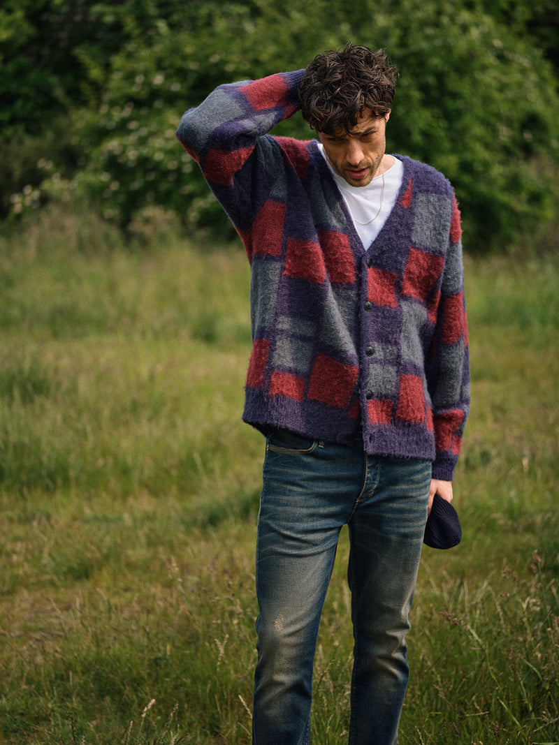 A person stands in a grassy field wearing the Blue de Gênes Block Knit Cardigan - Navy Pattern over a white T-shirt and blue jeans, with one hand on their head while looking down. Trees can be seen in the background.