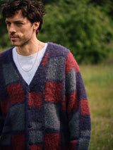 A man wears the Blue de Gênes Block Knit Cardigan in a navy pattern over a white t-shirt and thin necklace, standing outdoors with blurred greenery in the background.