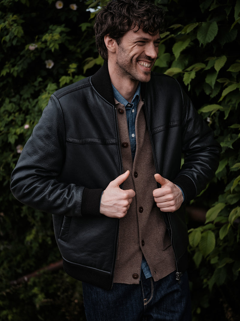 A man with curly hair smiles outdoors in front of green foliage, wearing a Blue de Gênes Gifu Knit Cardigan in Chocolate under a black leather jacket, paired with a blue shirt and jeans.