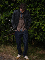 A man stands on grass by leafy bushes, looking down with his hands in his pockets. He wears the Blue de Gênes Rufo Shearling Jacket in Scarab over a brown cardigan, blue denim shirt, blue jeans, and white sneakers with tan accents.