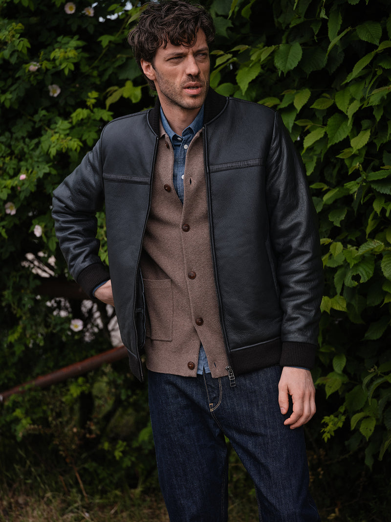 A man stands outdoors before lush foliage, wearing the Rufo Shearling Jacket - Scarab by Blue de Gênes over a brown cardigan, blue denim shirt, and dark jeans. He gazes thoughtfully to the side.