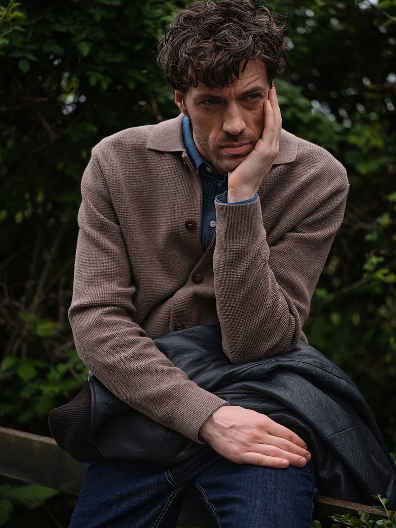 A man with curly hair sits outdoors, resting his face on one hand, looking thoughtful. He wears the Blue de Gênes Gifu Knit Cardigan in Chocolate over a denim shirt, with a black jacket on his lap and green foliage behind him.