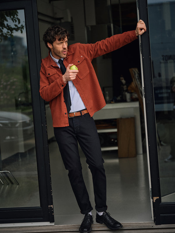 A man in a rust-orange Nagano Overshirt - Henna by Blue de Gênes, dress shirt, and tie leans against a door frame eating a green apple, standing casually with one foot crossed and looking to the side.