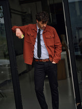A man stands in a doorway wearing a rust jacket over a striped shirt and the Nekutaki Tie #A - Navy from Blue de Gênes, paired with black pants and a brown belt. He leans casually, one hand in his pocket, looking down.