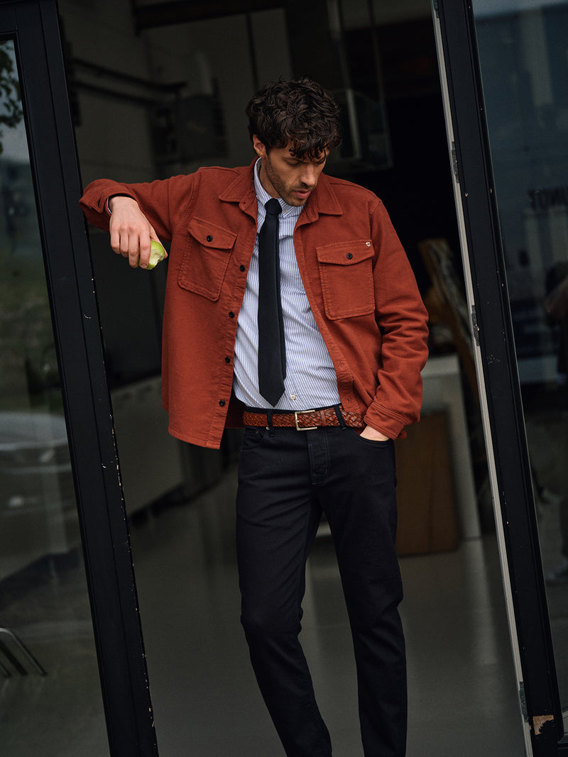 A man stands in a doorway wearing a rust jacket over a striped shirt and the Nekutaki Tie #A - Navy from Blue de Gênes, paired with black pants and a brown belt. He leans casually, one hand in his pocket, looking down.