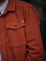 A close-up of a person wearing the Blue de Gênes Nagano Overshirt in Henna Moleskin cotton, with a flap pocket, black button, and embroidered shield logo above the pocket, layered over a blue and white striped shirt.