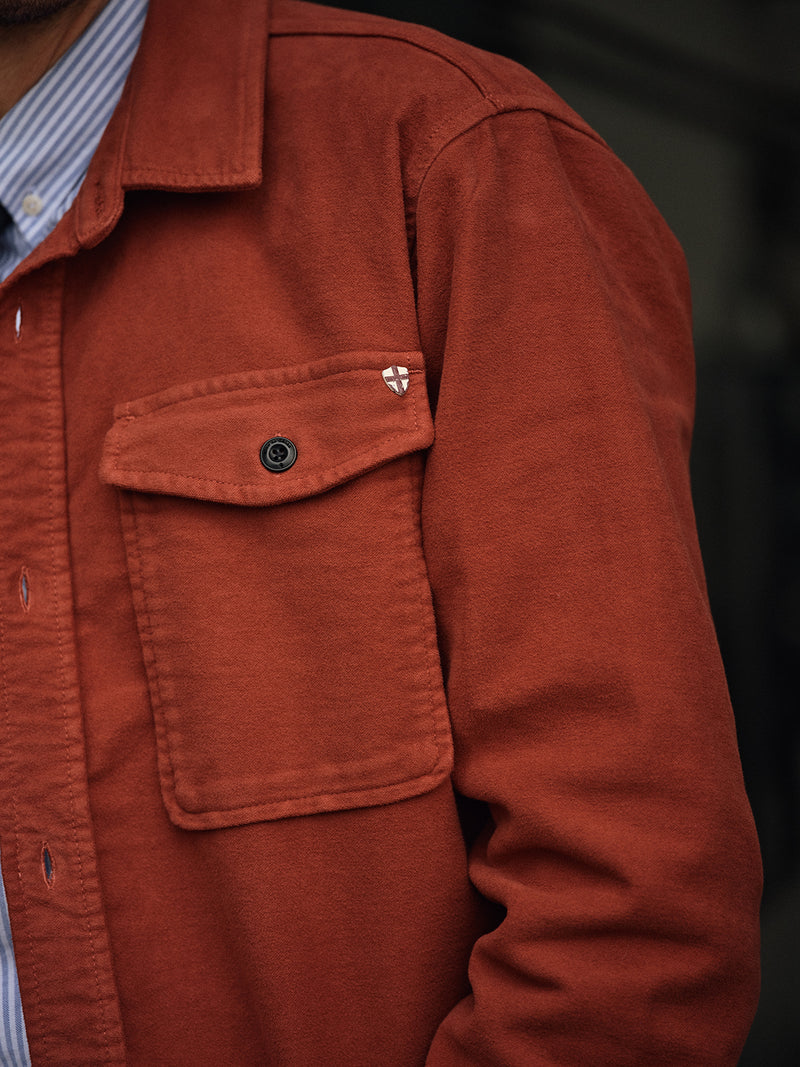 A close-up of a person wearing the Blue de Gênes Nagano Overshirt in Henna Moleskin cotton, with a flap pocket, black button, and embroidered shield logo above the pocket, layered over a blue and white striped shirt.