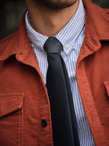 A person wears a Blue de Gênes Nagano Overshirt in Henna over a blue and white striped dress shirt with a dark, textured necktie. The photo is cropped to display only the chest and neck area.