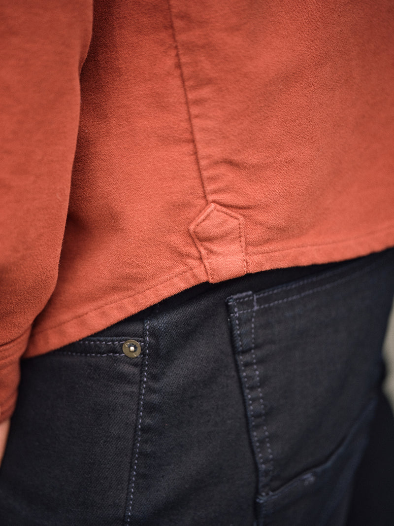 Close-up of a person wearing the Nagano Overshirt in Henna by Blue de Gênes with dark blue jeans, highlighting the shirts side detail, hem, and the jeans’ back pocket.