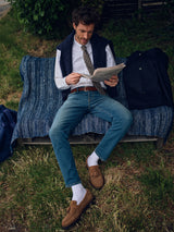 A man, dressed in a white shirt, tie, dark vest, Blue de Gênes Vinci Topaz Vintage Jeans - Used, white socks, and brown loafers, reads a newspaper on a blue bench. A dark coat lies beside him among grass and bushes.