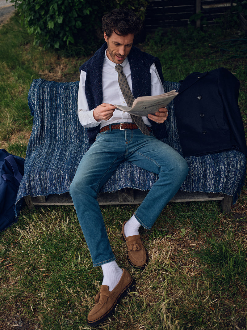 A man, dressed in a white shirt, tie, dark vest, Blue de Gênes Vinci Topaz Vintage Jeans - Used, white socks, and brown loafers, reads a newspaper on a blue bench. A dark coat lies beside him among grass and bushes.
