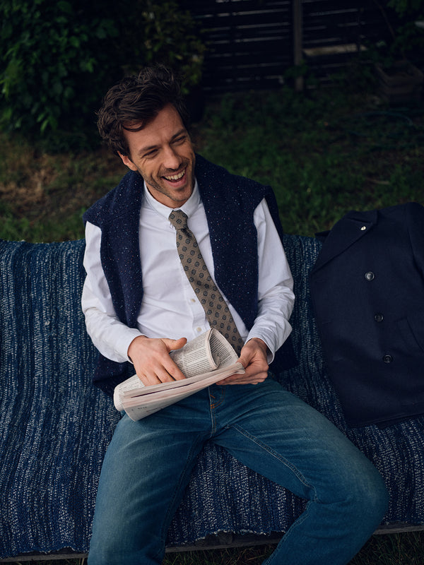 A smiling man with tousled hair sits on a blue bench outdoors, reading a newspaper while wearing the Blue de Gênes Honshu Crewneck Knit in Navy draped over his shoulders, paired with a white shirt, patterned tie, and jeans. A coat lies beside him.