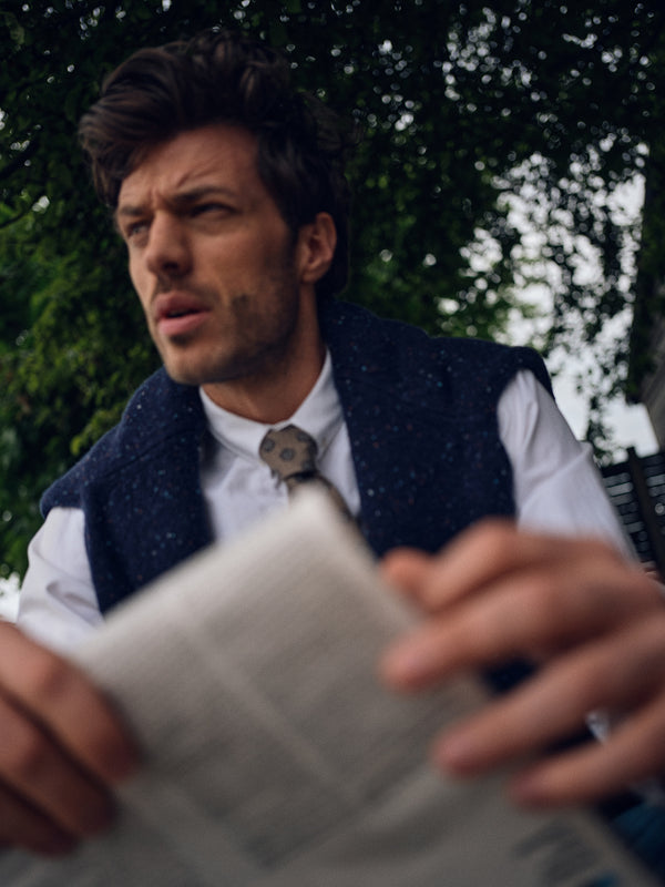 A man in the Blue de Gênes Honshu Crewneck Knit in navy sits outdoors with a newspaper, looking thoughtfully to the side. Lush green foliage forms a blurred background.