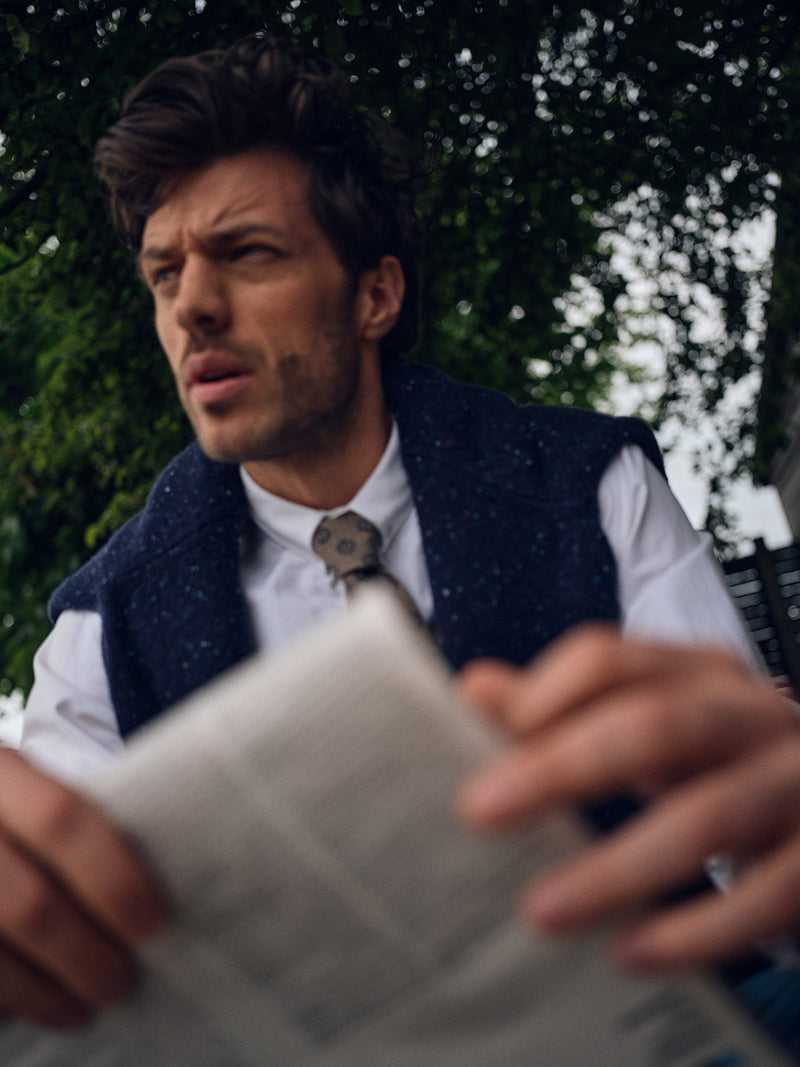 A man in the Blue de Gênes Honshu Crewneck Knit in navy sits outdoors with a newspaper, looking thoughtfully to the side. Lush green foliage forms a blurred background.