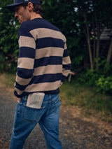 Wearing the Blue de Gênes Toyama Stripe Knit L/S in Dark Navy with blue jeans and a cap, a person walks outdoors, a folded newspaper in their back pocket, surrounded by green foliage and a gravel path.