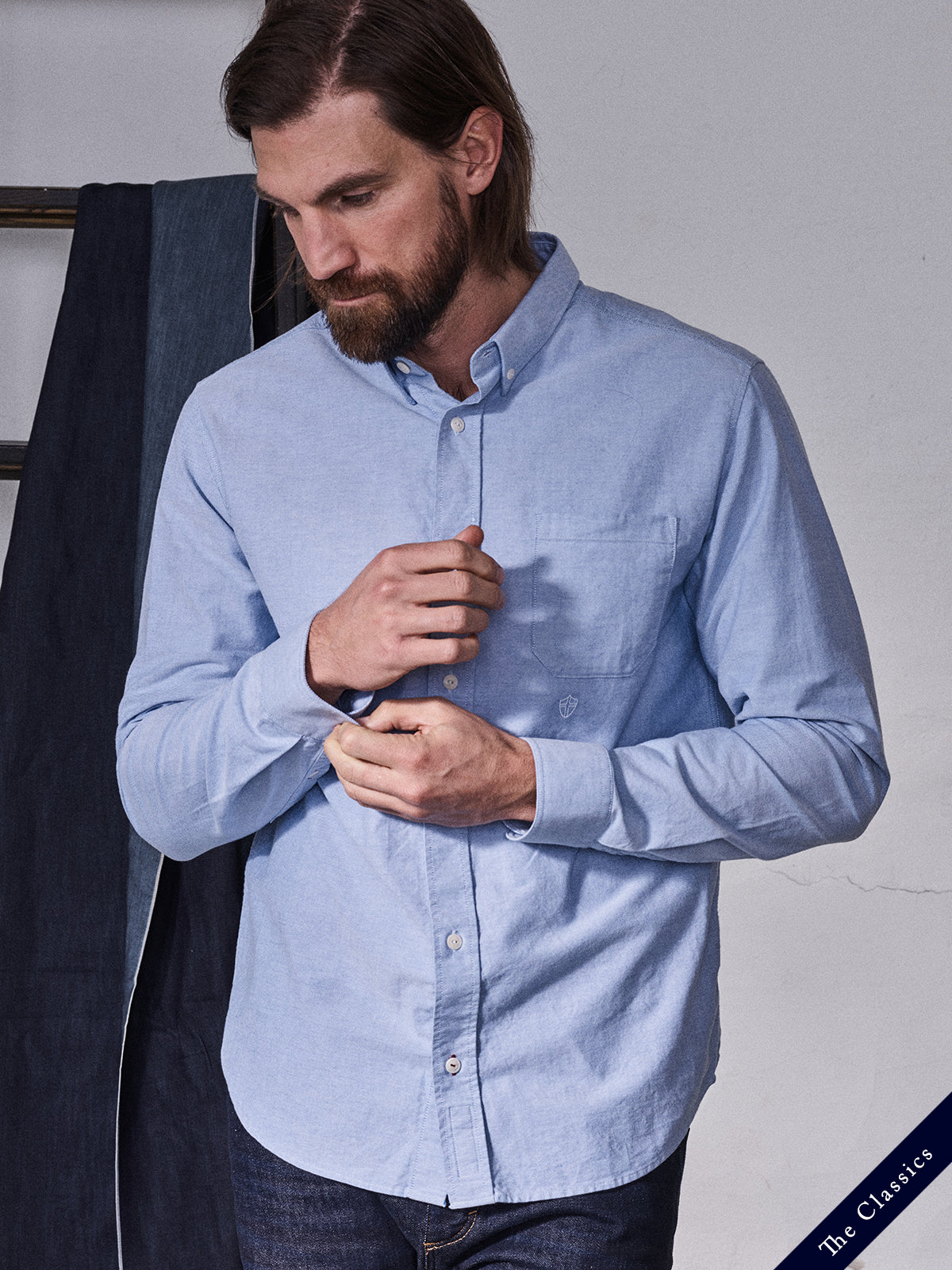 A man with shoulder-length brown hair and a beard fastens a cuff while wearing the BGsandro Oxford Shirt - Blue by Blue de Gênes. He pairs it with dark jeans, framed by draped fabric and 
