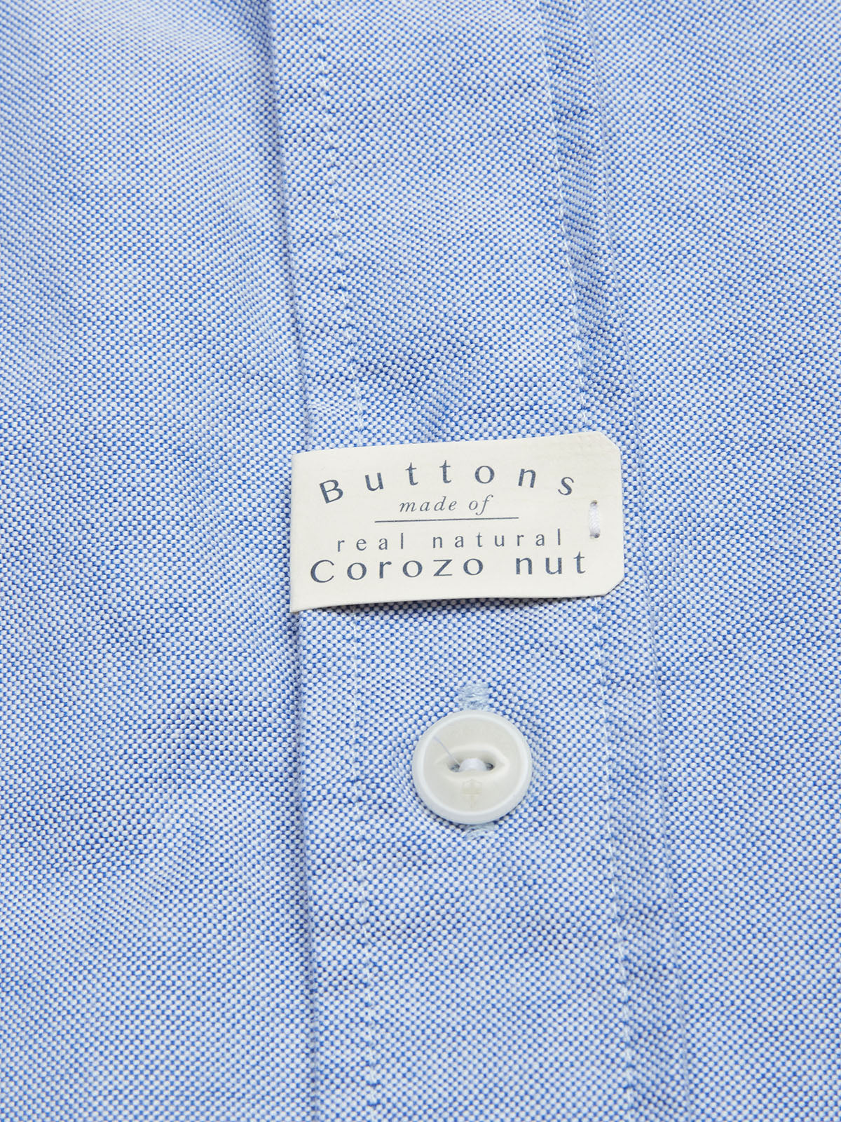 A close-up of the BGsandro Oxford Shirt - Blue by Blue de Gênes, crafted from textured organic cotton, with a tag above a white button that states, 